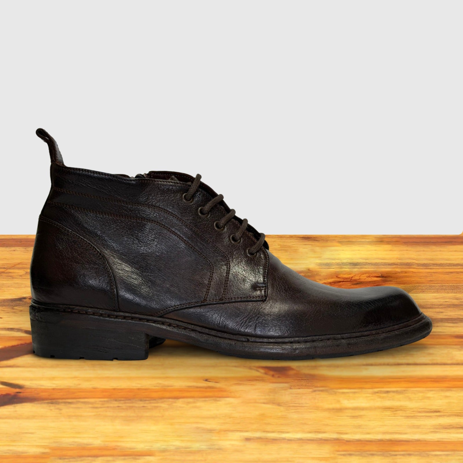 A dark brown leather lace-up ankle boot with a low stacked heel, shown in profile on a wooden surface against a light gray background.