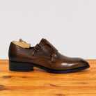 A chestnut leather double strap monk shoe with brogue detailing and a black heel, displayed on a wooden surface against a light gray background.