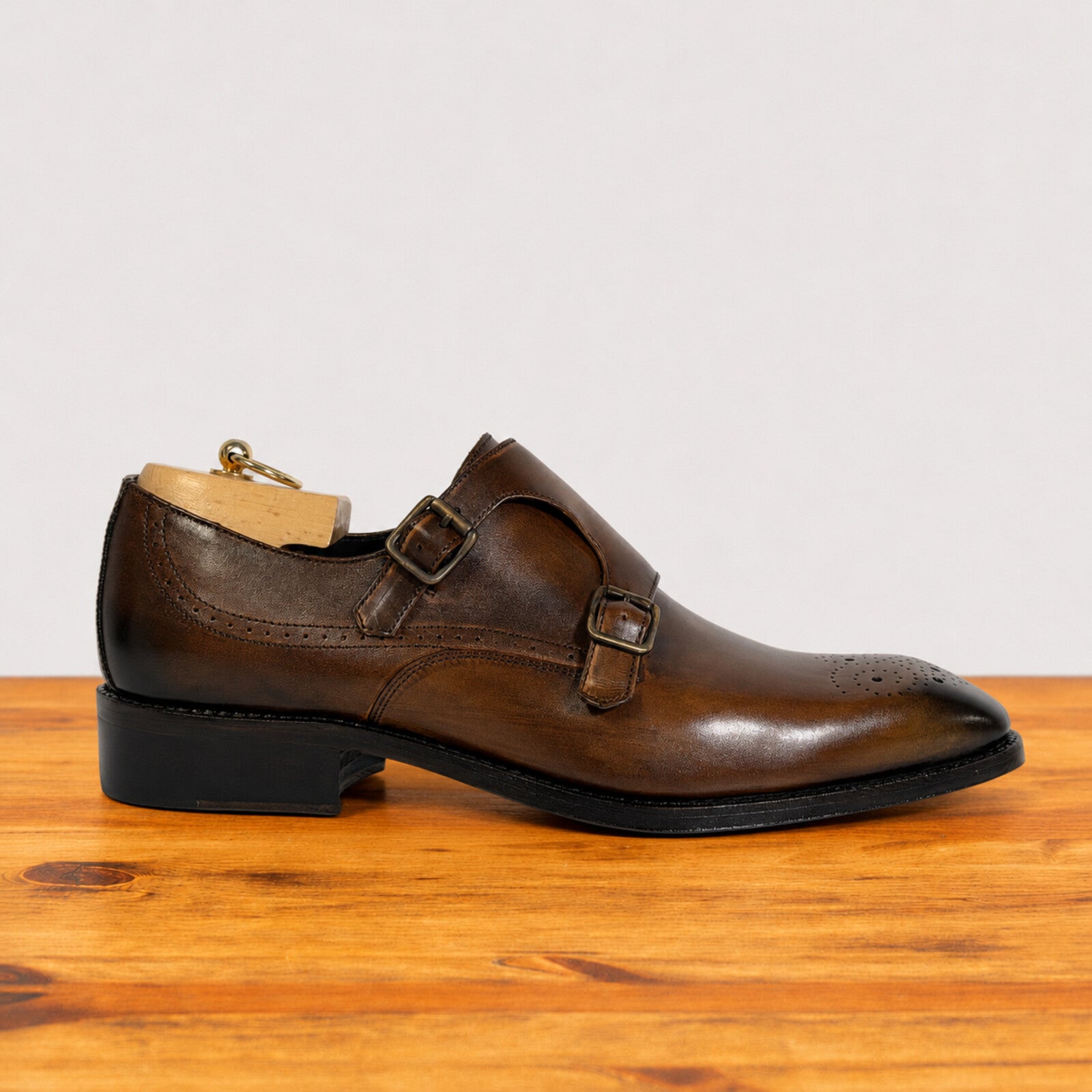 A chestnut leather double strap monk shoe with brogue detailing and a black heel, displayed on a wooden surface against a light gray background.