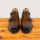 A pair of chestnut leather double monk shoes with brogue detailing on the toes, displayed on wooden shoe trees on a wooden surface against a light gray background.