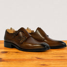 A pair of chestnut leather double monk shoes with brogue detailing and black soles, displayed on a wooden surface against a light gray background.