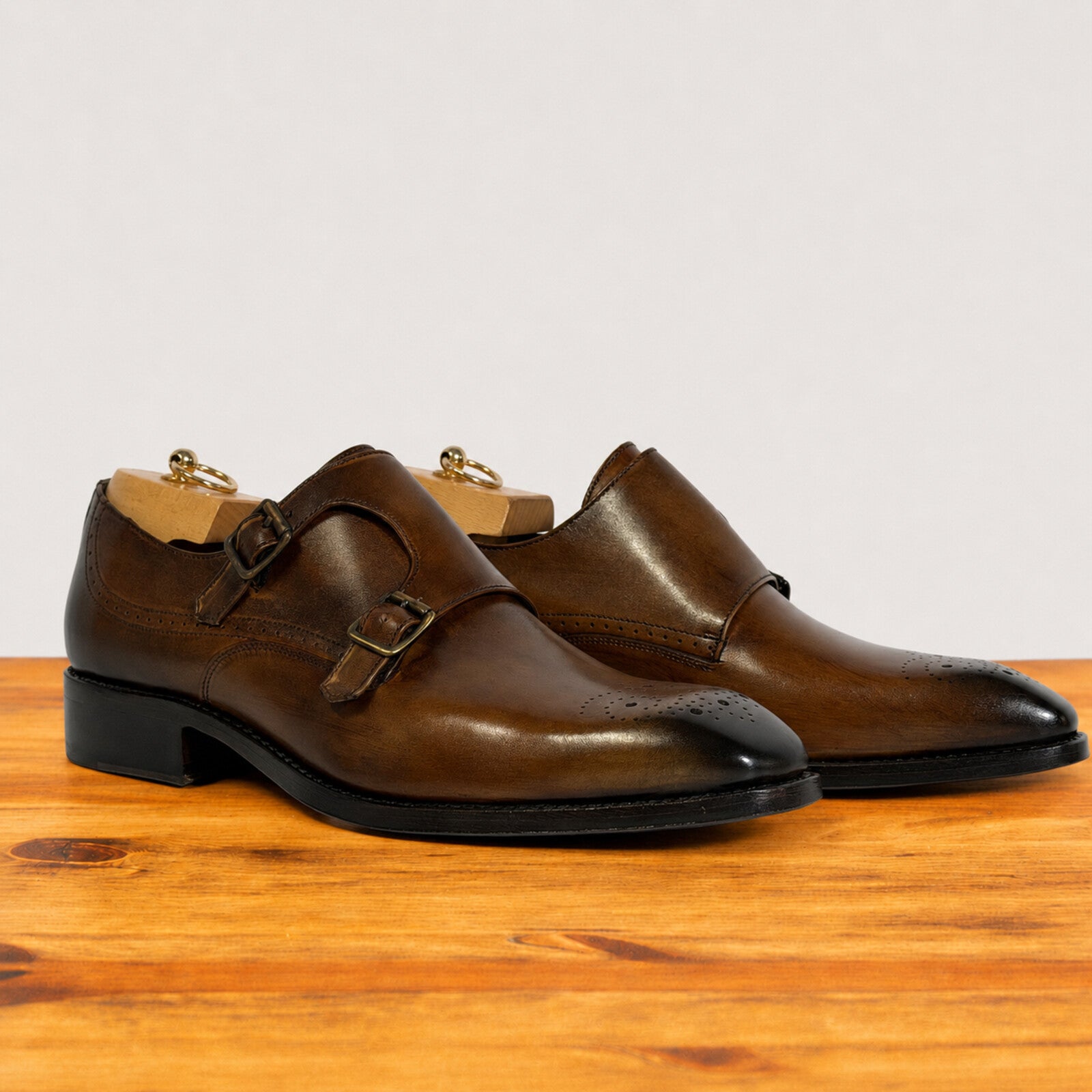 A pair of chestnut leather double monk shoes with brogue detailing and black soles, displayed on a wooden surface against a light gray background.