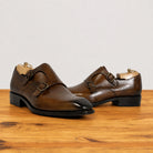 A pair of chestnut leather double monk shoes with brogue detailing and brass-tone buckles, displayed on a wooden surface against a light gray background.