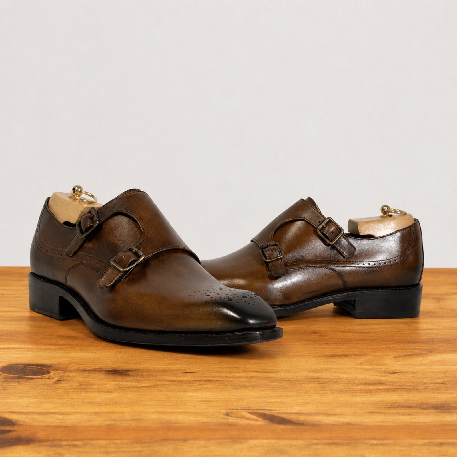 A pair of chestnut leather double monk shoes with brogue detailing and brass-tone buckles, displayed on a wooden surface against a light gray background.