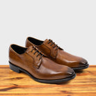 A Pair of brown, wholecut blucher dress shoe with a lace-up closure, displayed on a wooden surface.