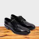 A Pair of black, wholecut blucher dress shoe with a lace-up closure, displayed on a wooden surface.