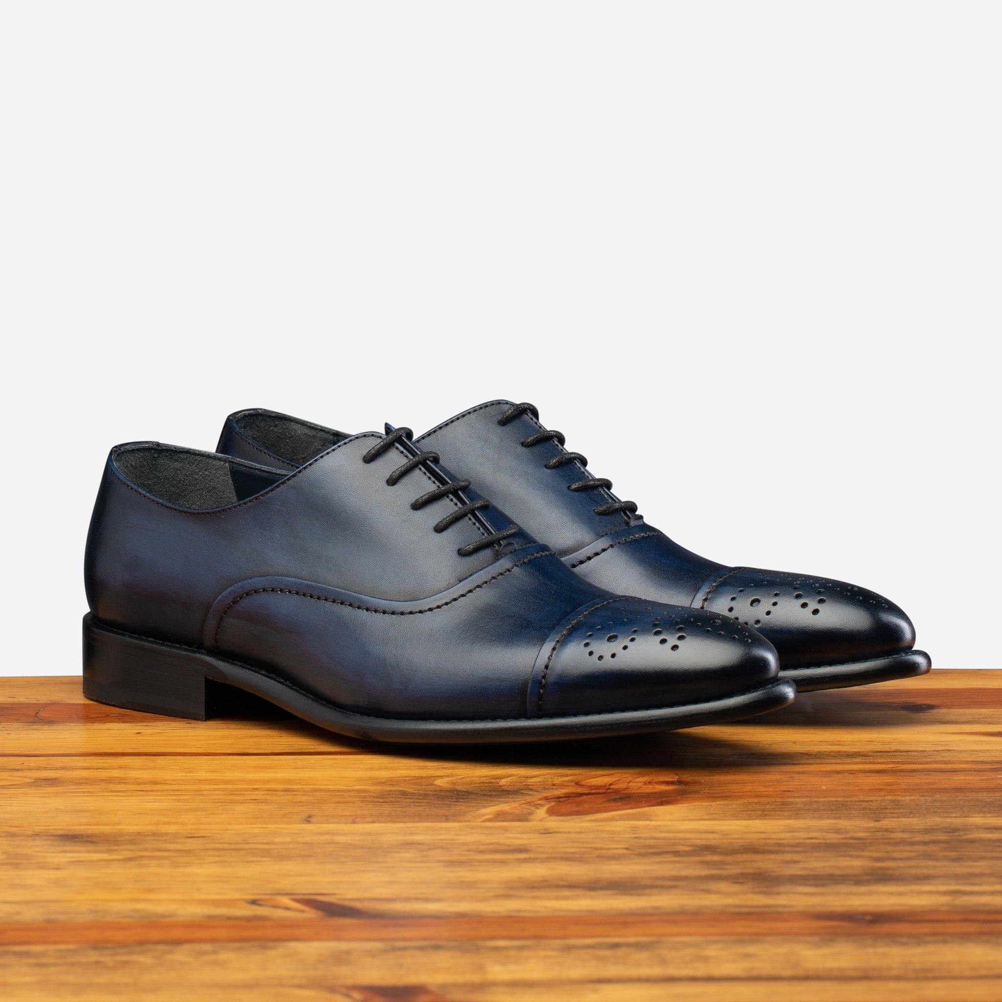 Pair of a dark blue leather dress shoe toe featuring decorative perforations and stitching, resting on a wooden surface.