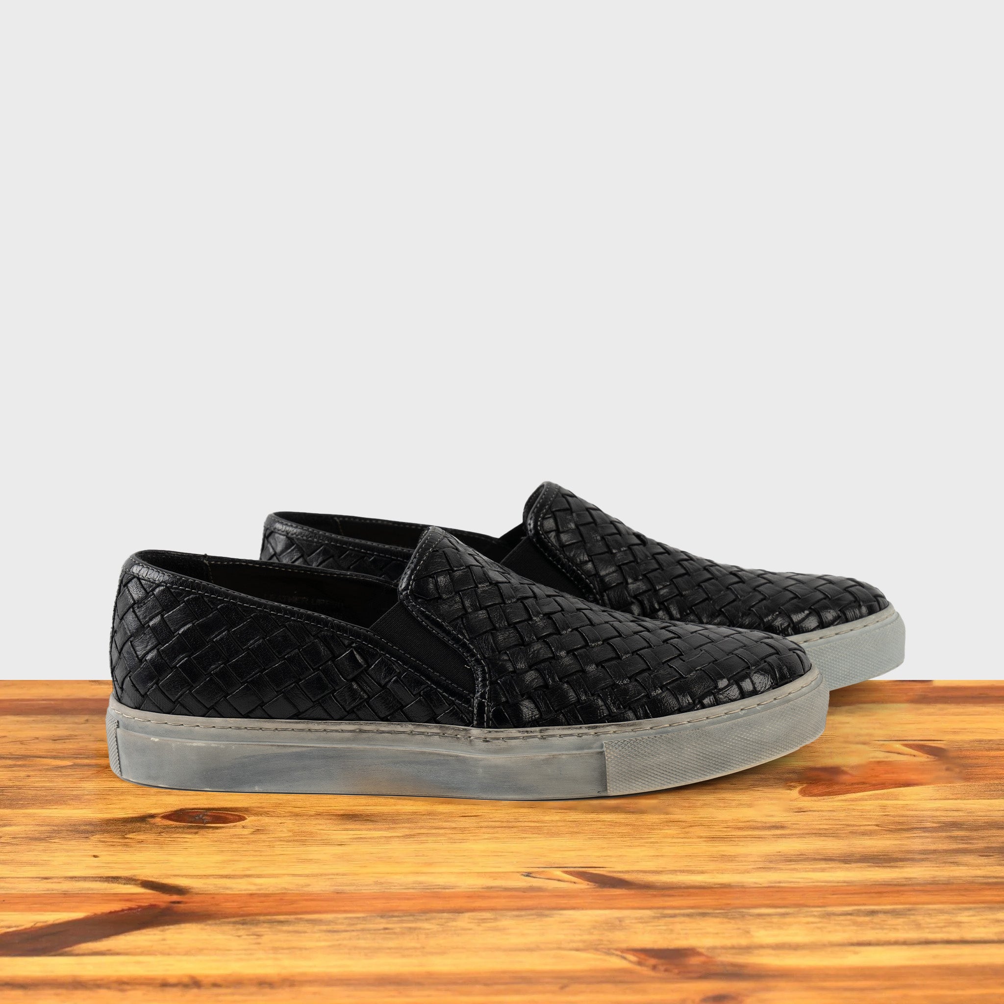 A Pair of black woven slip-on shoe on a wooden surface with a white background
