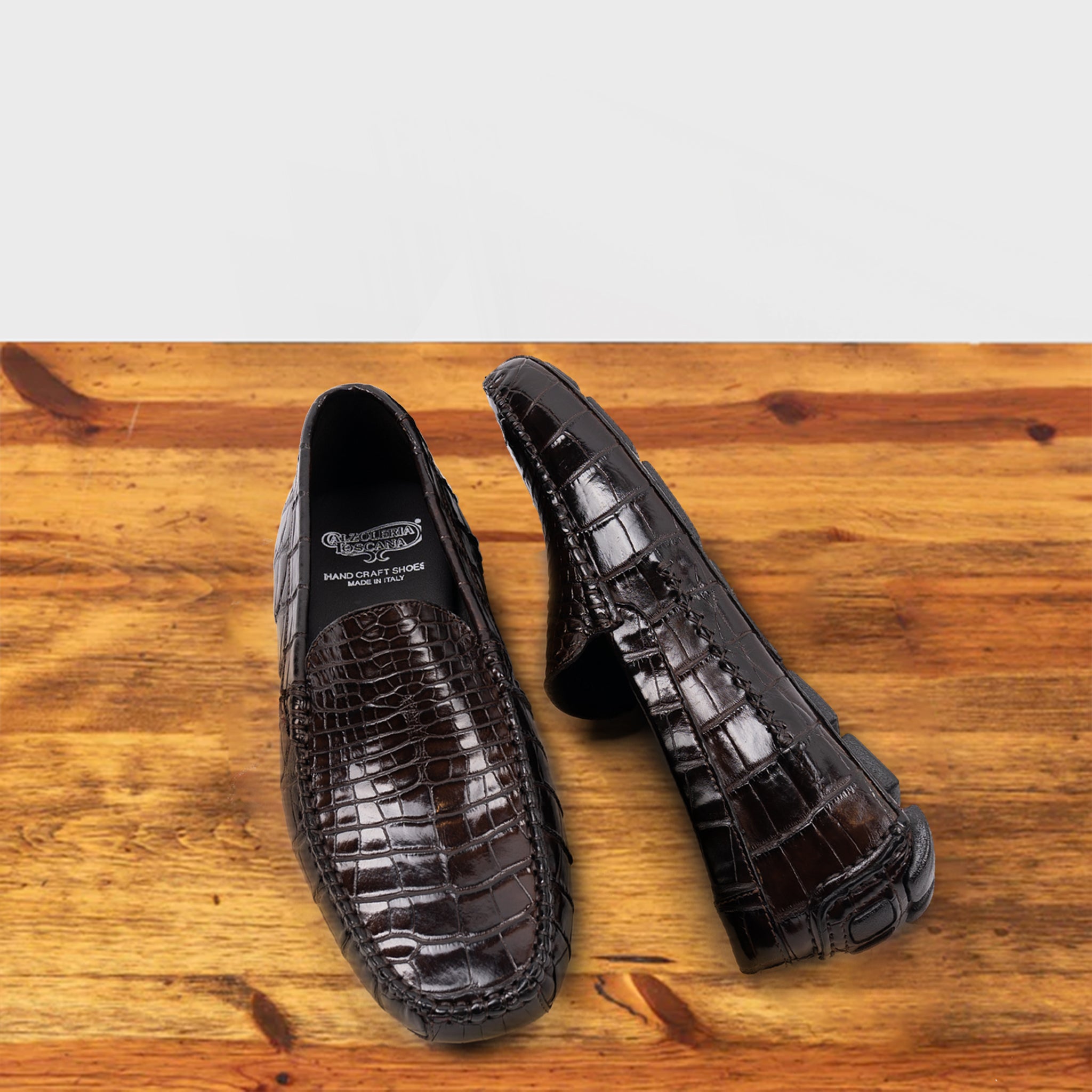A Pair of Dark Brown crocodile-patterned loafer on a wooden surface with a light gray background