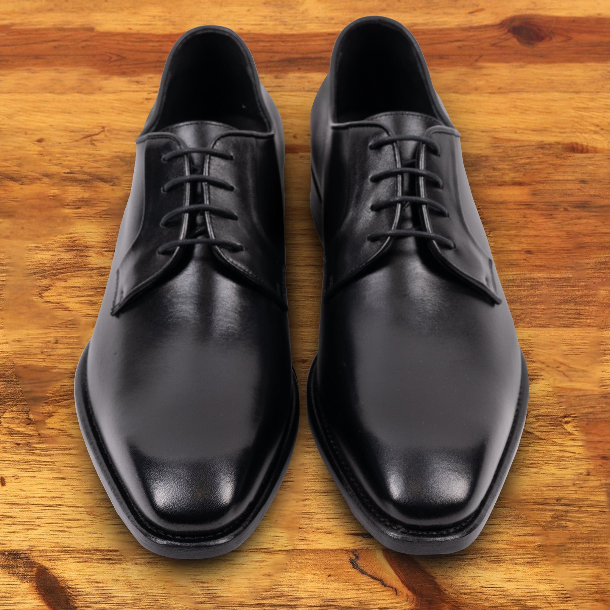 A Pair of black, wholecut blucher dress shoe with a lace-up closure, displayed on a wooden surface.