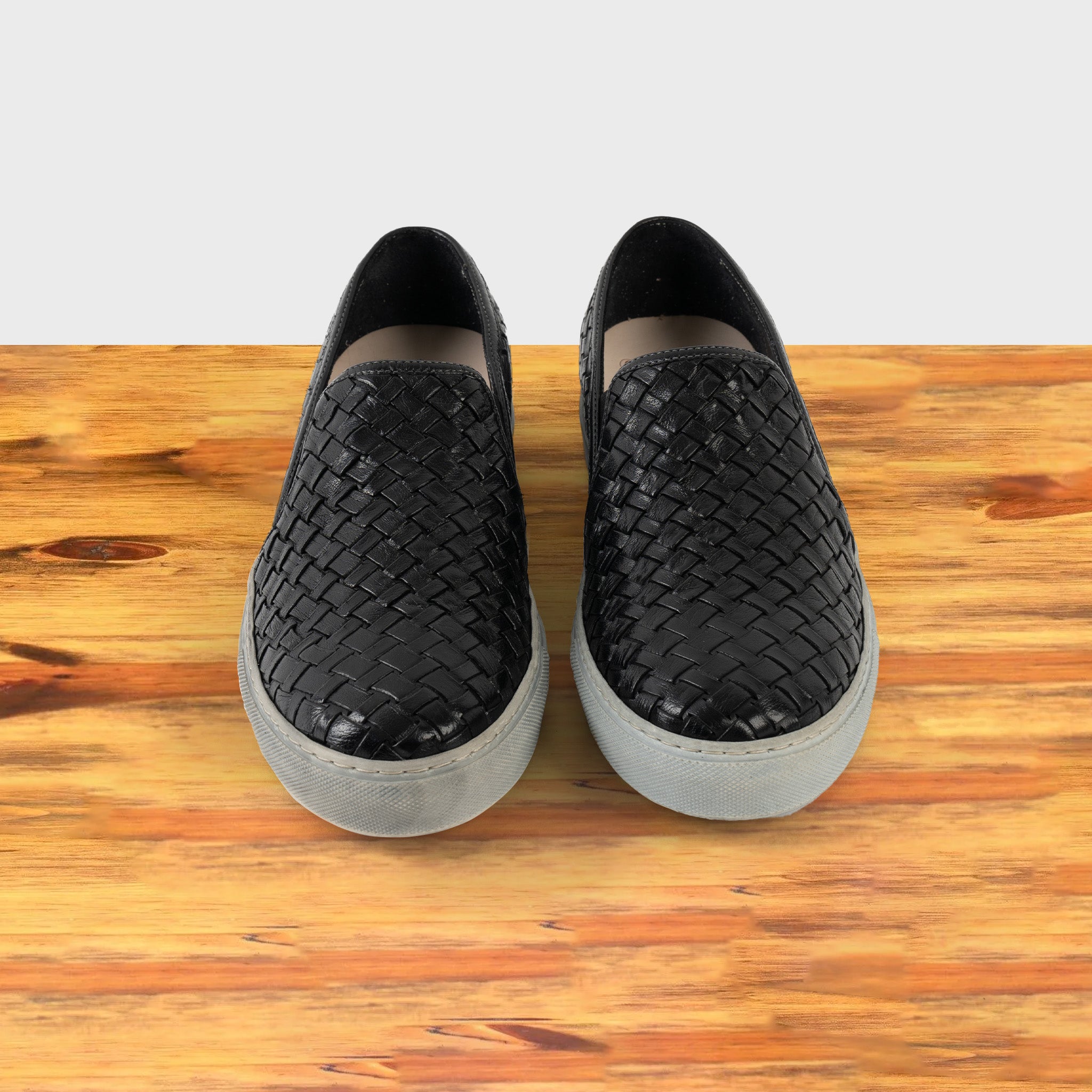 A Pair of black woven slip-on shoe on a wooden surface with a white background