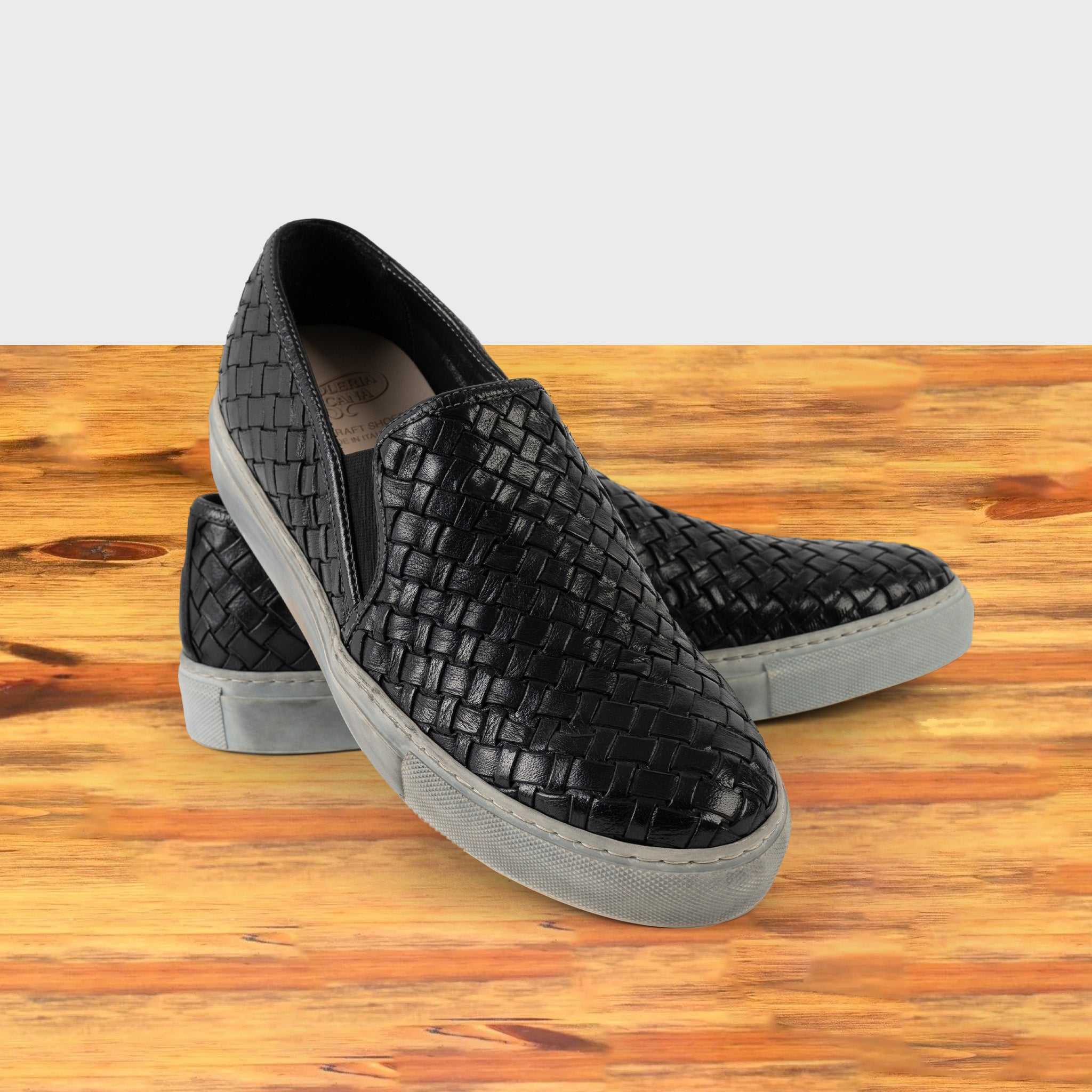 A Pair of black woven slip-on shoe on a wooden surface with a white background