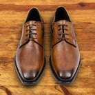 A Pair of brown, wholecut blucher dress shoe with a lace-up closure, displayed on a wooden surface.