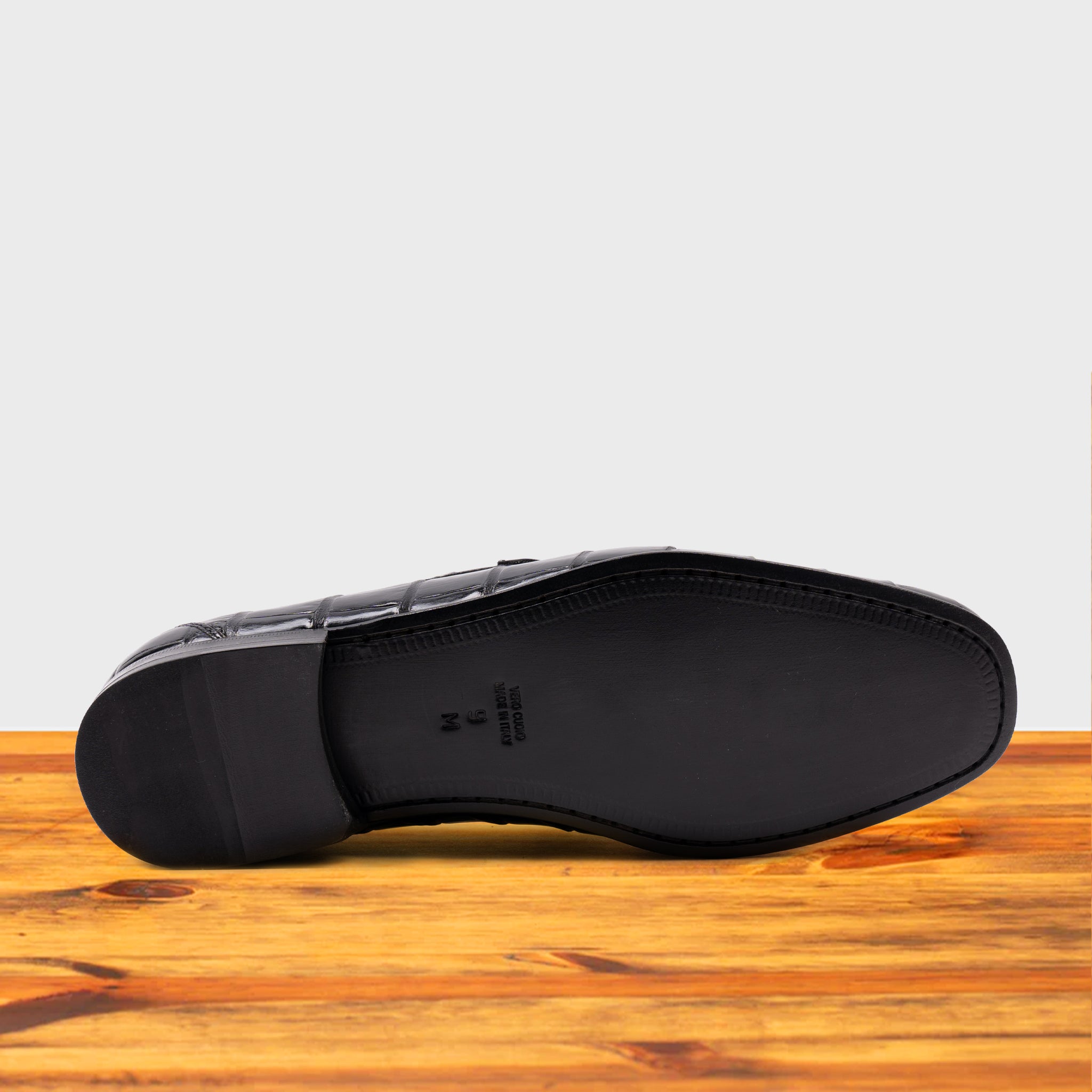 Outsole of Black crocodile-patterned loafer on a wooden surface with a light gray background