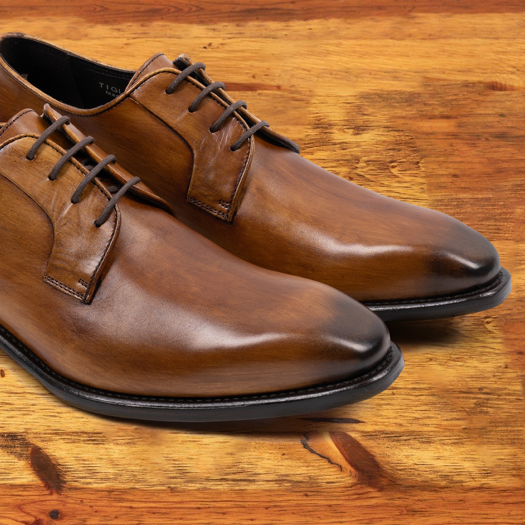 A Pair of brown, wholecut blucher dress shoe with a lace-up closure, displayed on a wooden surface.