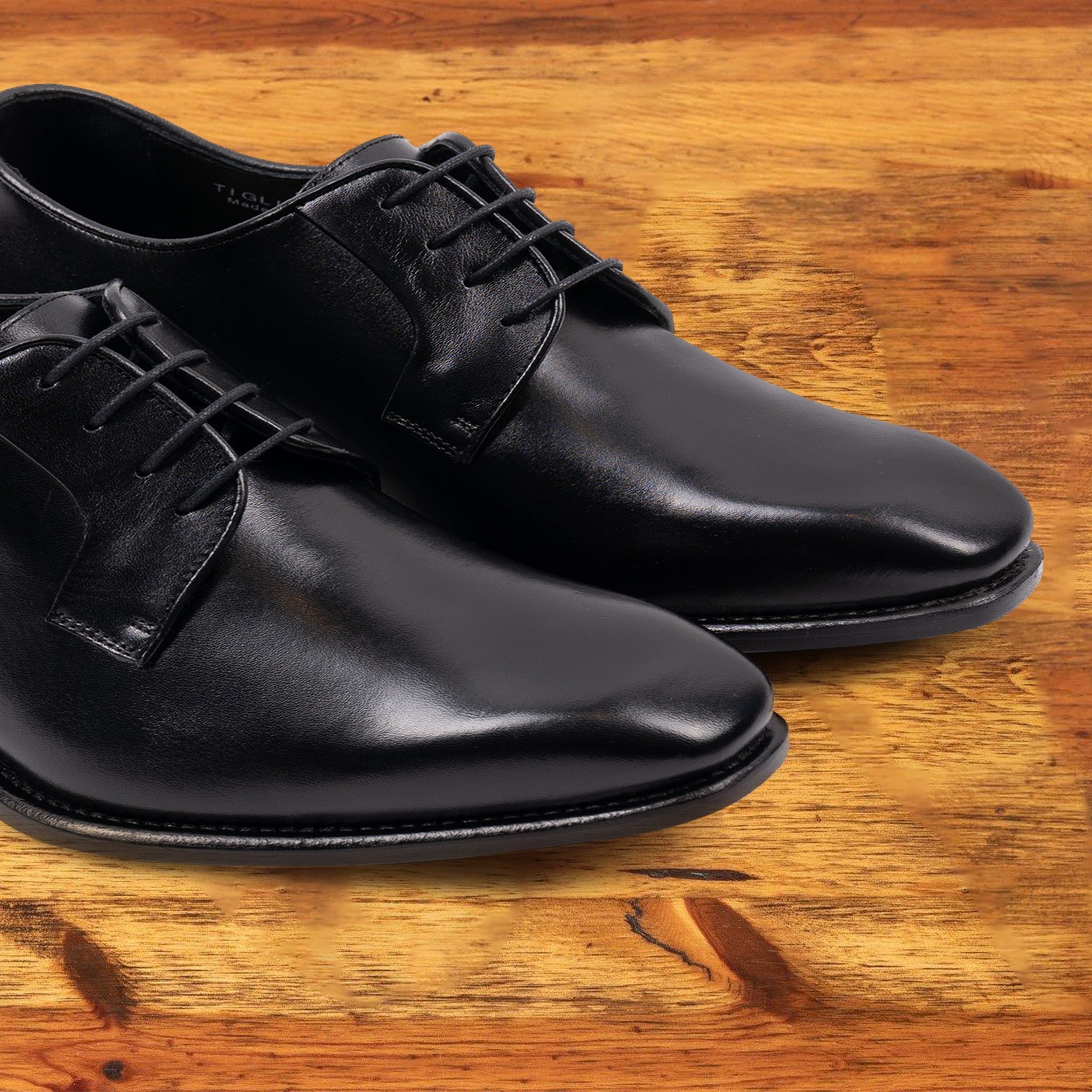 A Pair of black, wholecut blucher dress shoe with a lace-up closure, displayed on a wooden surface.