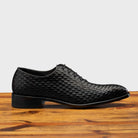 Black woven dress shoe on a wooden surface with a light gray background