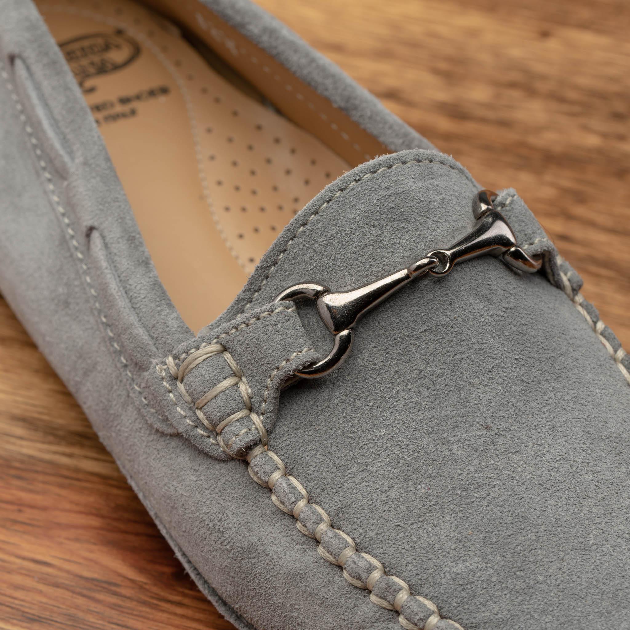 Silver velour suede driver shoes with a metallic bit, placed on a wooden surface.