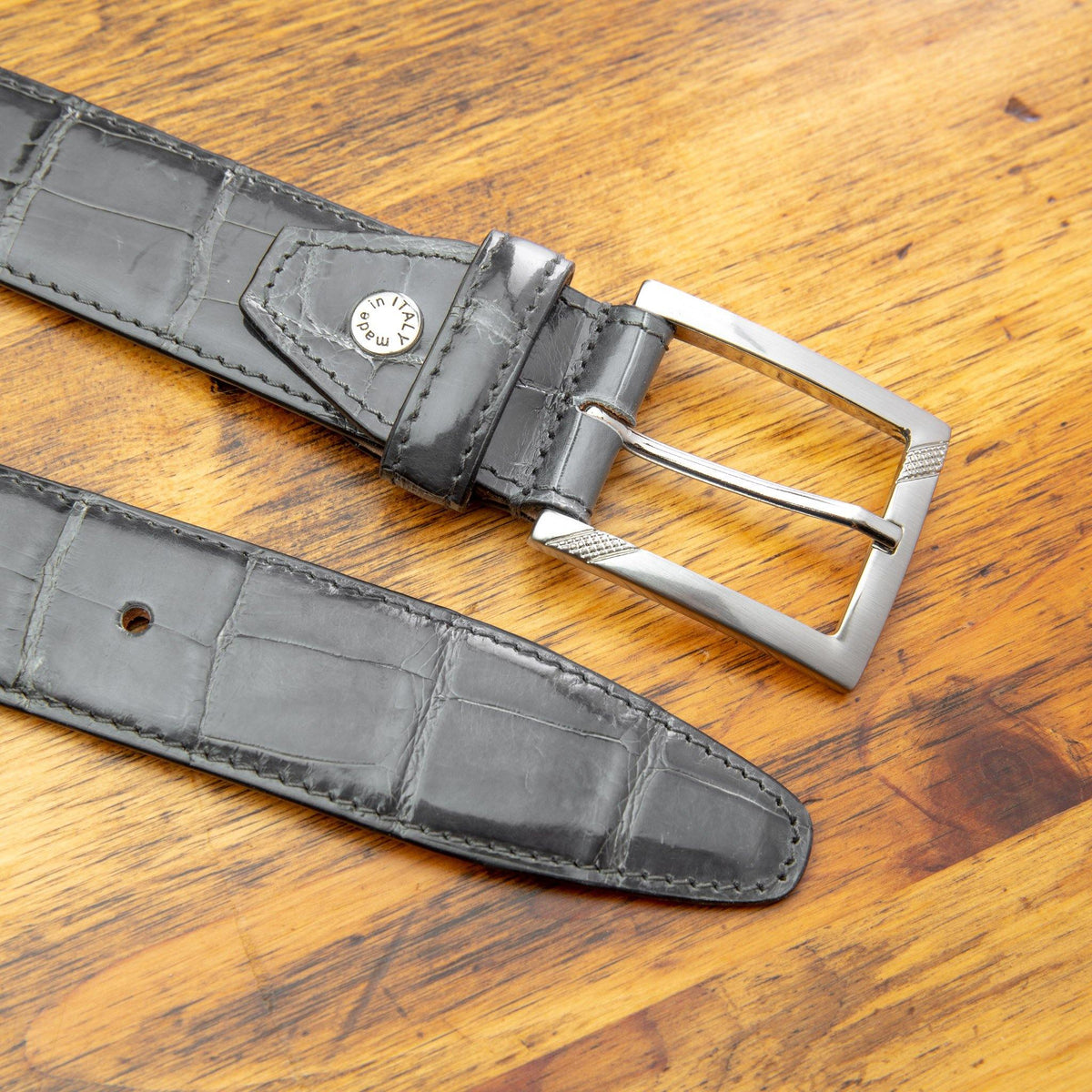 C8099 Gray crocodile leather belt with a polished silver buckle, coiled on a wooden surface