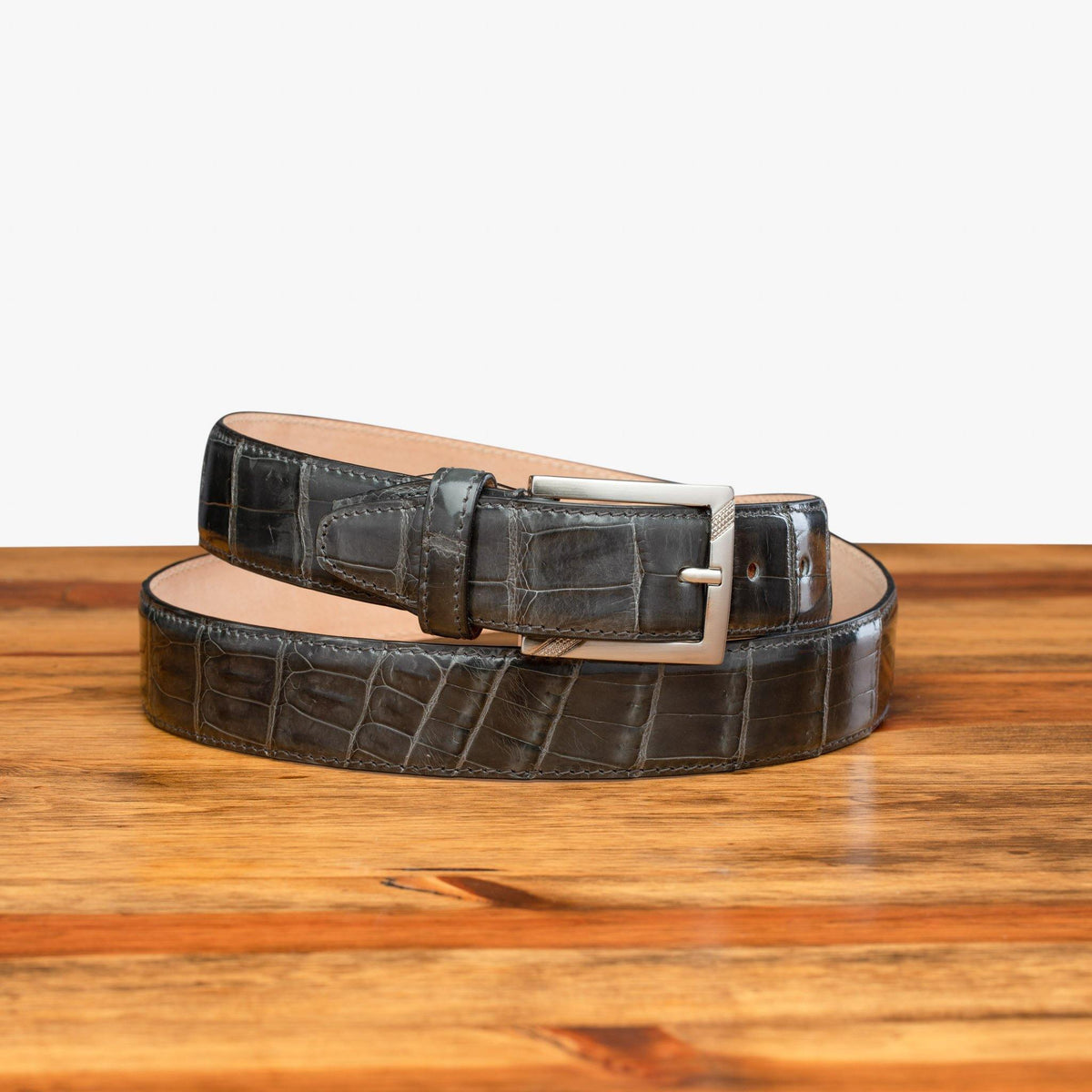 C8099 Gray crocodile leather belt with a polished silver buckle, coiled on a wooden surface