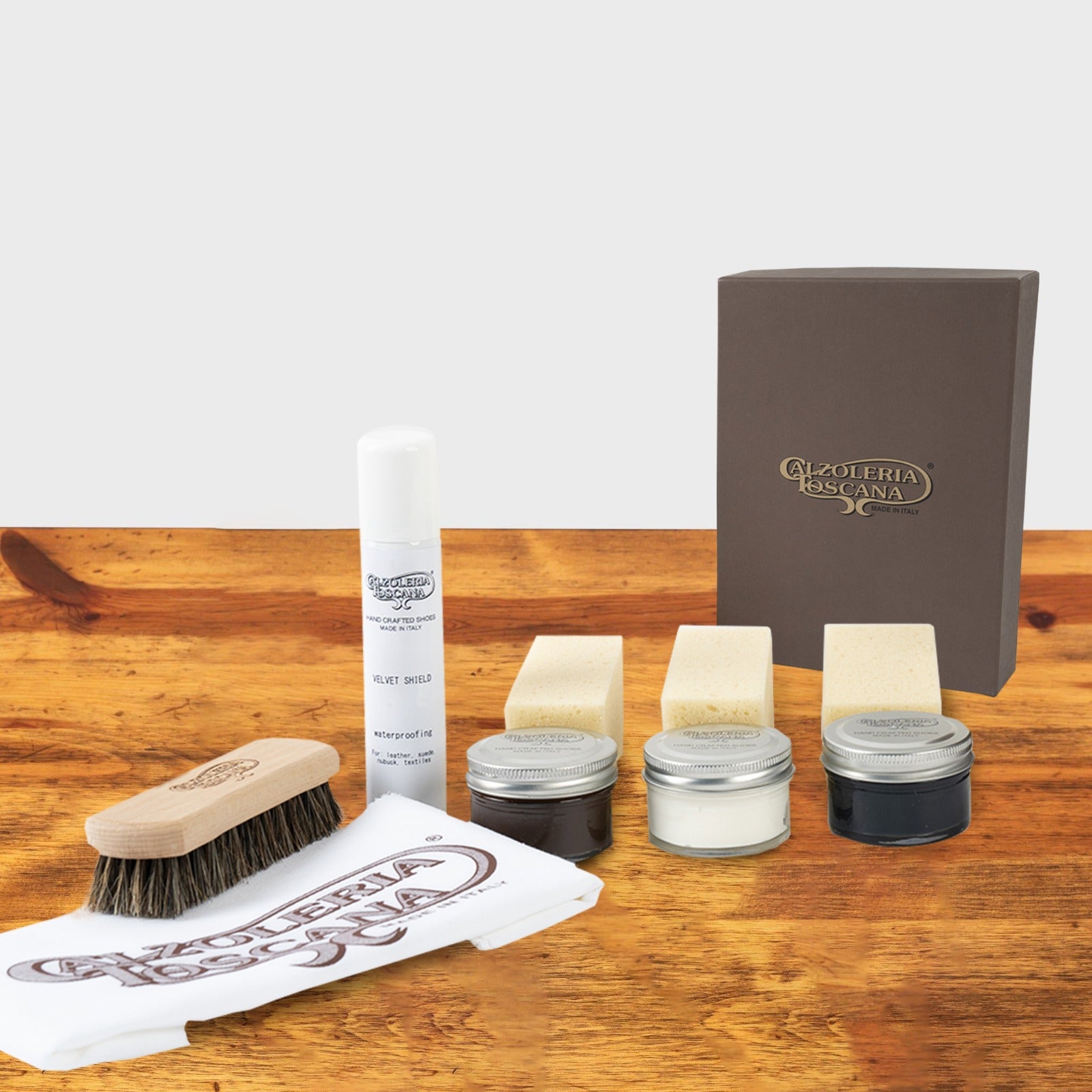 Shoe care kit, featuring sponges, polish tins, a waterproofing spray, and a wooden brush, arranged neatly on a wooden surface.