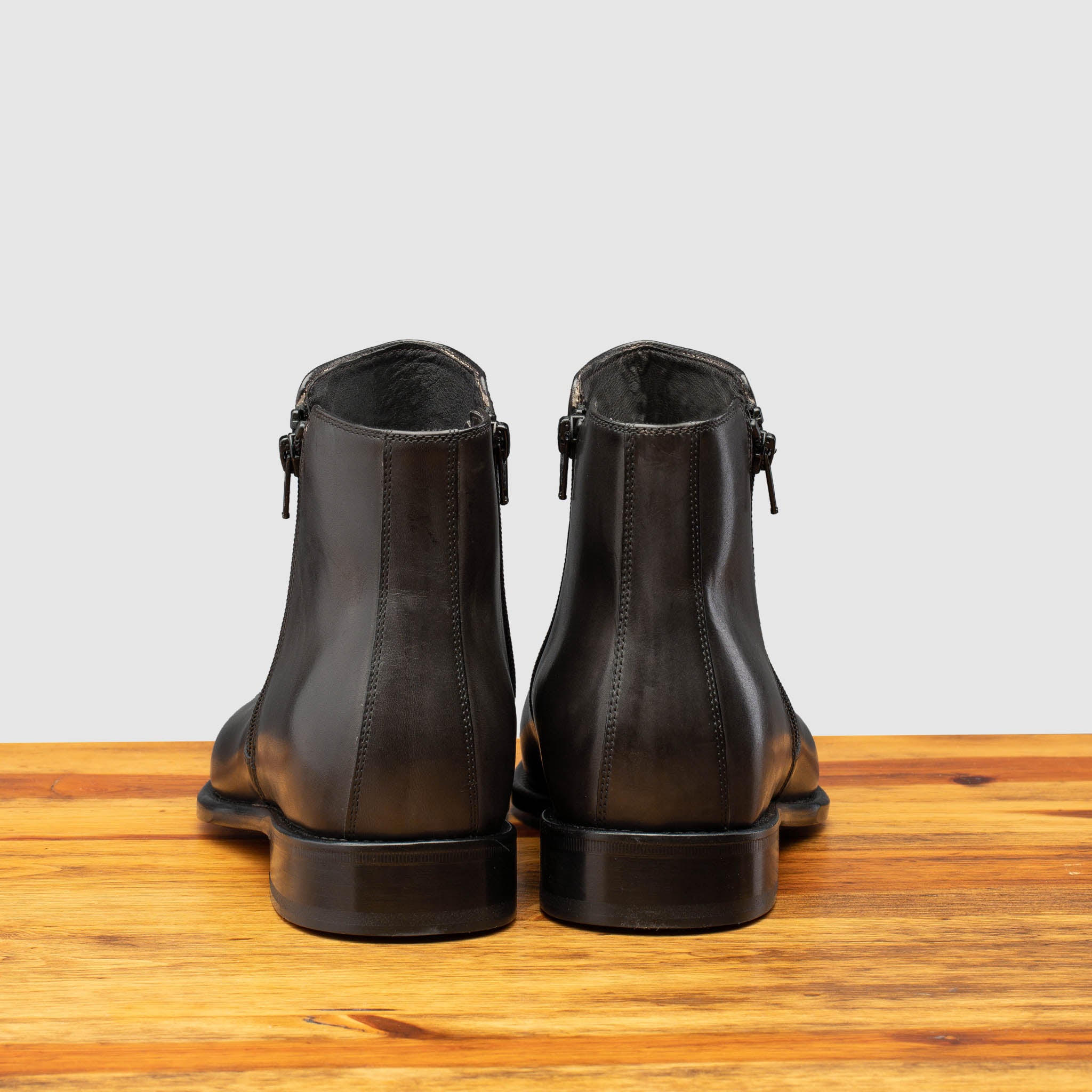 Back view of dark brown leather ankle boot with zipper on a wooden surface and light gray background