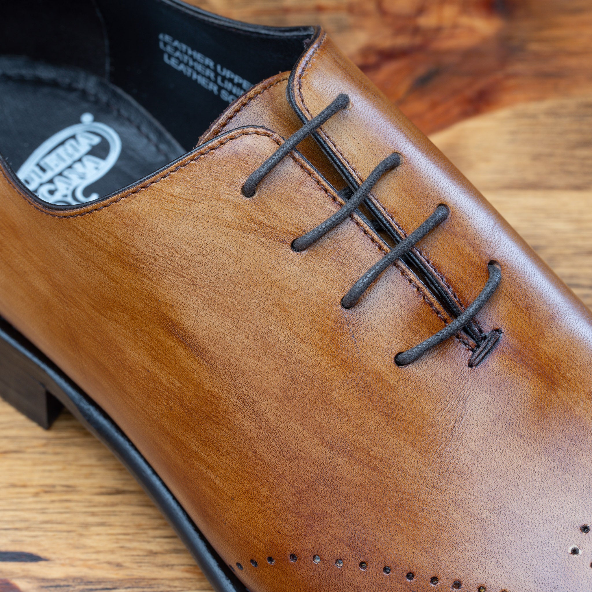 A chestnut wholecut leather Oxford shoe with a lace-up closure, displayed on a wooden surface.