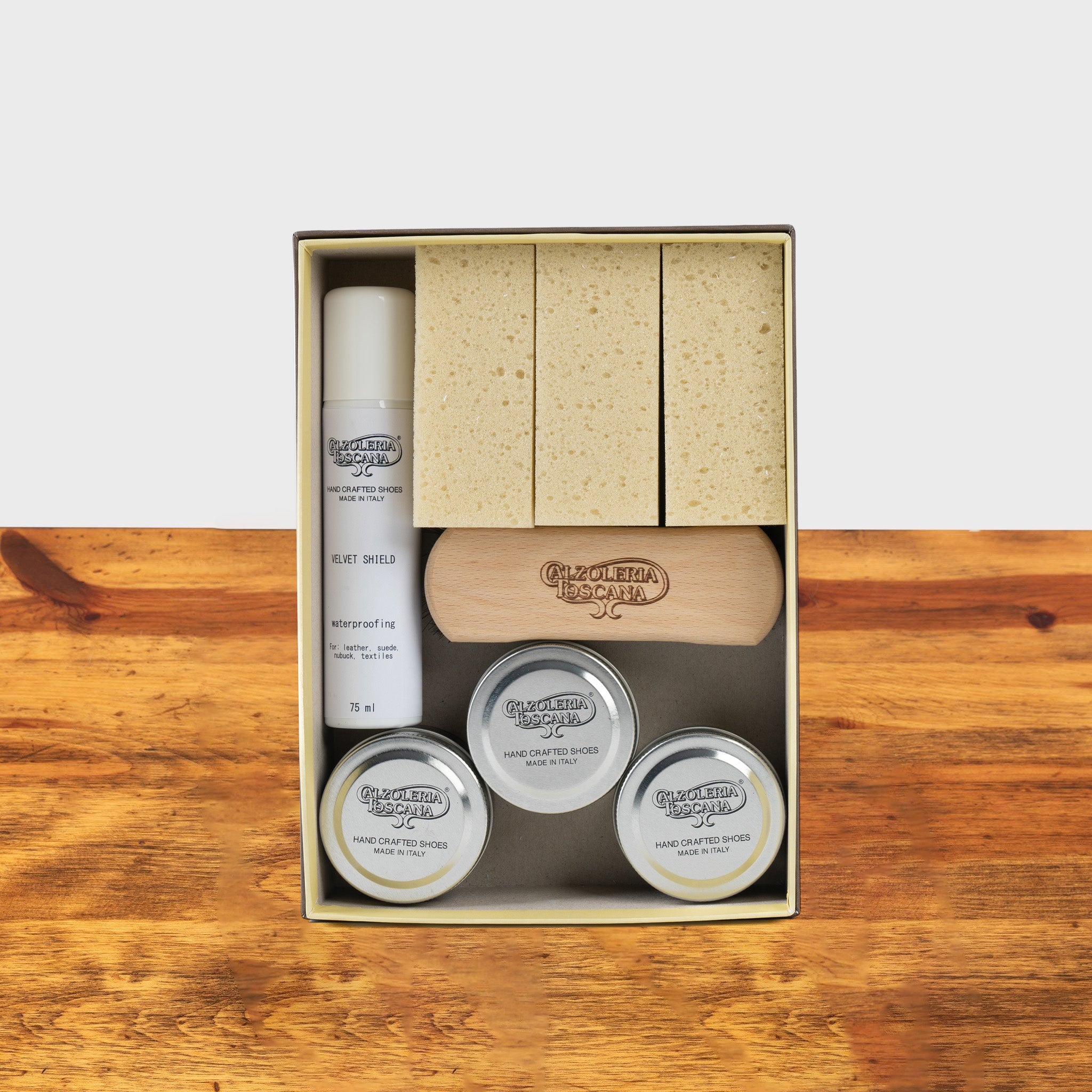 Shoe care kit in a box featuring sponges, polish tins, a waterproofing spray, and a wooden brush, arranged neatly on a wooden surface.